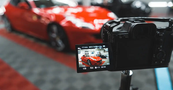 A camera on a tripod pointed toward a red sports car. The car is visible in focus on the camera’s monitor screen.
