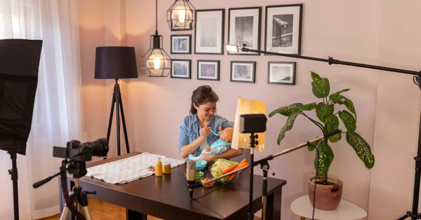 A woman sitting at a desk in front of a camera on a tripod. She is smiling and pretending to feed a baby doll.