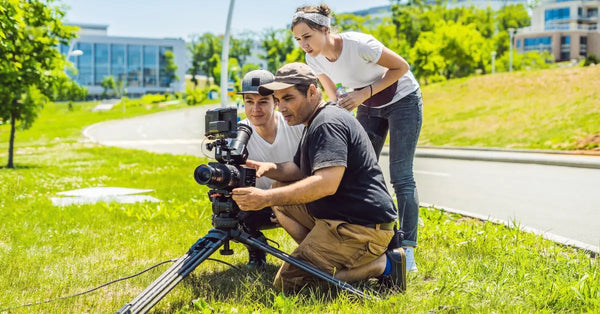 A camera operator and two other crew members looking into a camera on a tripod. They are outdoors on a sunny day.