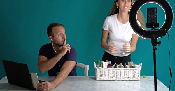 A woman faces a smartphone and ring light, filming a video. Next to her, a man at a laptop makes a “shush” gesture.