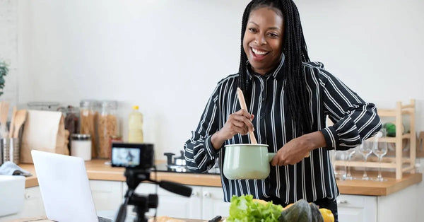 A woman stirring the contents of a small pot with a wooden spoon. She smiles and faces a camera on a tripod.