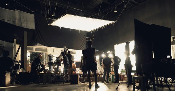 A film crew works on a set that is lit by multiple lights. Scrim diffusers are set up above and behind the set.