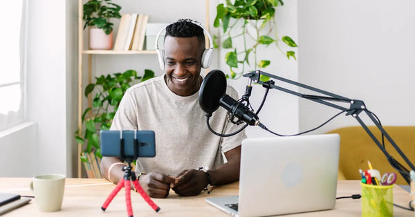 A young man smiles as he looks at a phone attached to a tabletop tripod. There’s a laptop and a microphone on the desk.