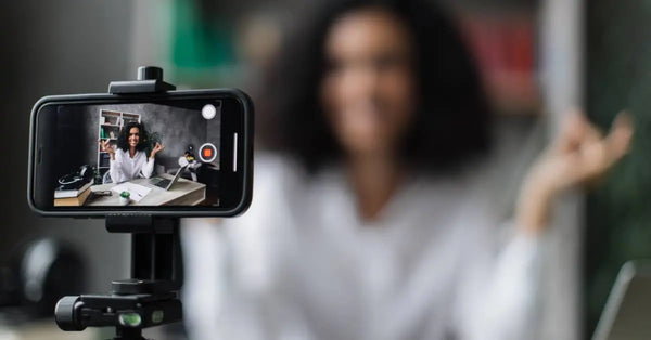 A woman in a white blouse smiles and sits at a desk. A smartphone affixed to a tripod records her actions.