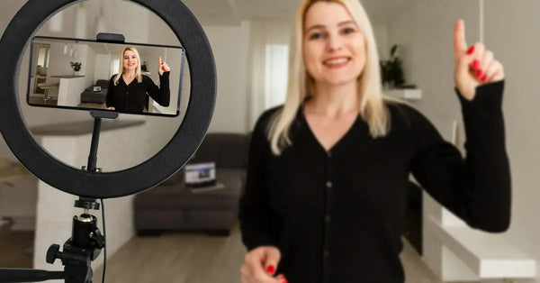 A young woman in a black shirt smiles and gestures as she faces a smartphone and ring light on a tripod.