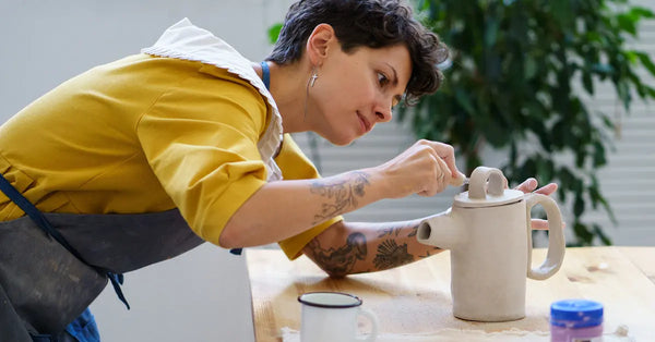 A short-haired woman in an apron crouches as she works on a ceramic jug. A smartphone on a tripod records her.